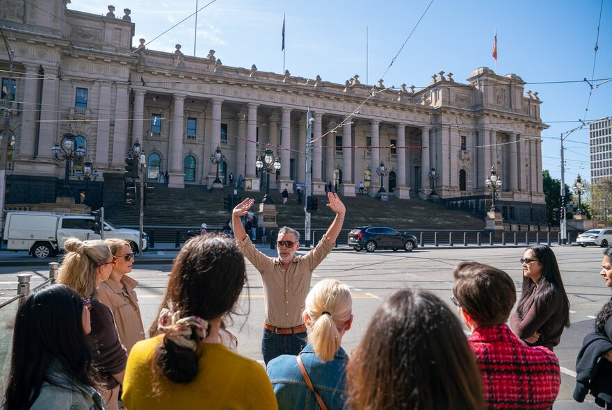 The back of a group of people on a walking tour in front of a group leader with his hands in the air at Parliament House. 