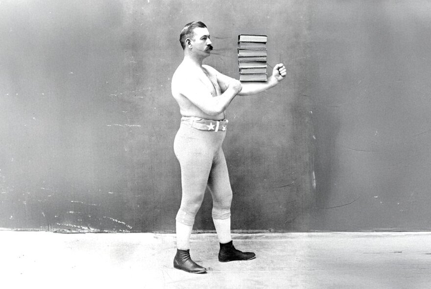 An early black and white image of a man with a thick moustache in a boxing pose with books balanced on his arm. 