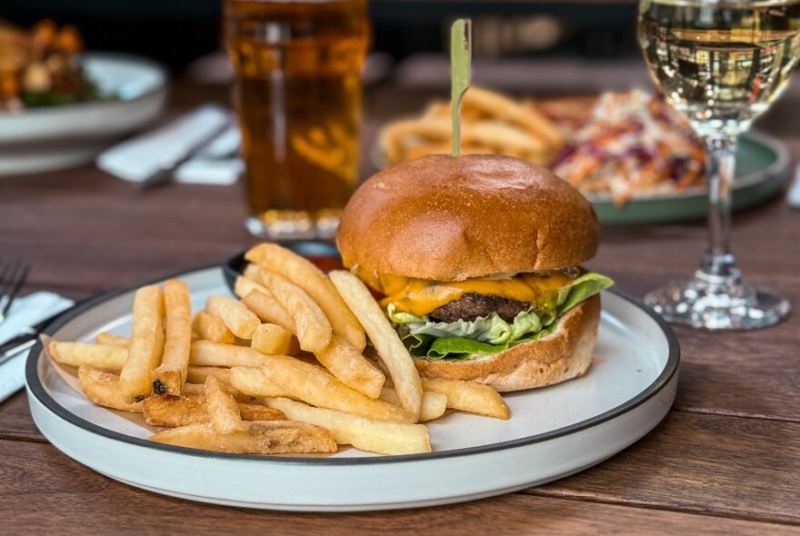 A hamburger and fries on a plate on a wooden table, with a pot of beer and a glass of wine.