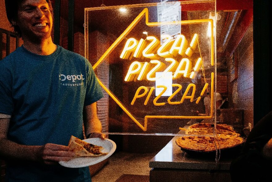 A smiling tour guide in front of a shop that has the words PIZZA PIZZA PIZZA written on the window, and holding a slice of pizza in his hands.