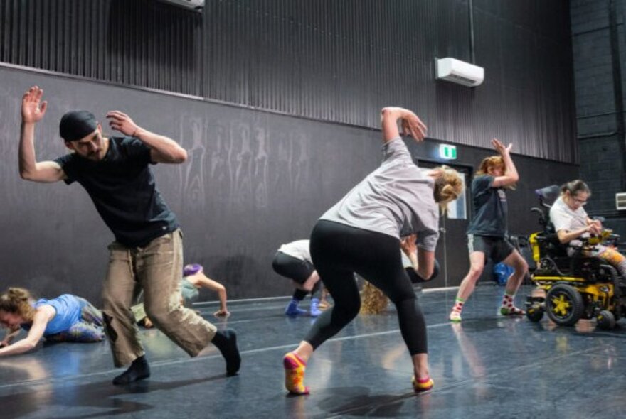 A studio space with black walls and a dark floor, with people moving around in the space, one of them lying on the ground.