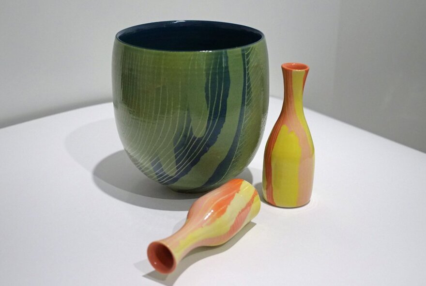 A green ceramic vase and two smaller, narrow-necked orange and yellow vases, one lying on its side, on a white table. 