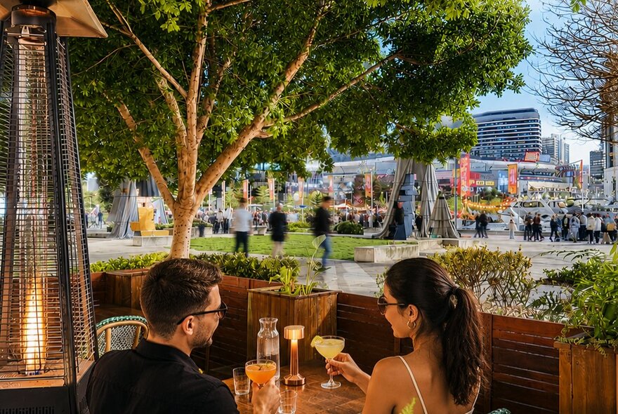 Guests dining under shaded trees outside in a cafe courtyard, with views of the Docklands precinct, pedestrians and buildings in the background.