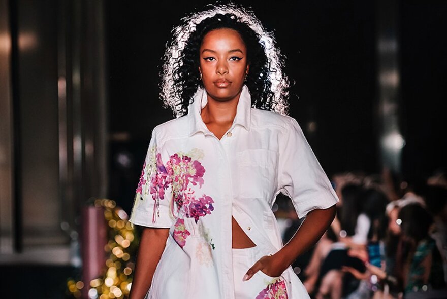 Fashion model walking the runway with a seated audience in the background.