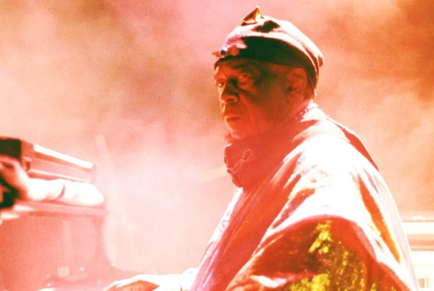 A man in a glittering gold jacket, playing the piano on a smokey stage.