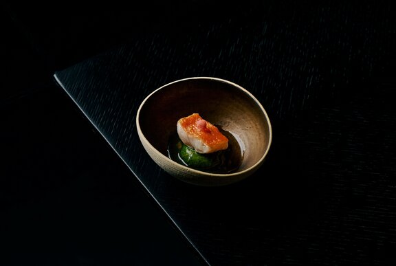A single piece of seared kingfish in a small Japanese bowl, resting on a green leaf surrounded by a dark sauce.