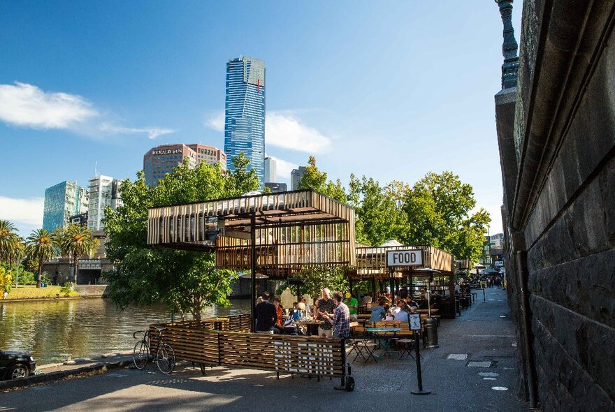 The wooden outdoor seating area of Pilgrim Bar along the banks of the Yarra River, with city buildings and trees in the background.