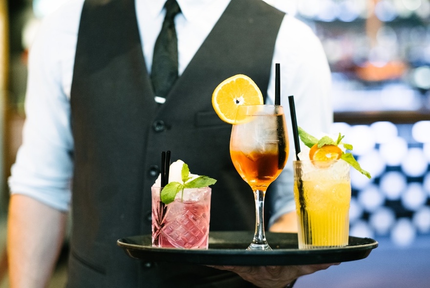 Waiter in a black vest, white shirt and black tie, carrying a tray of cocktails.