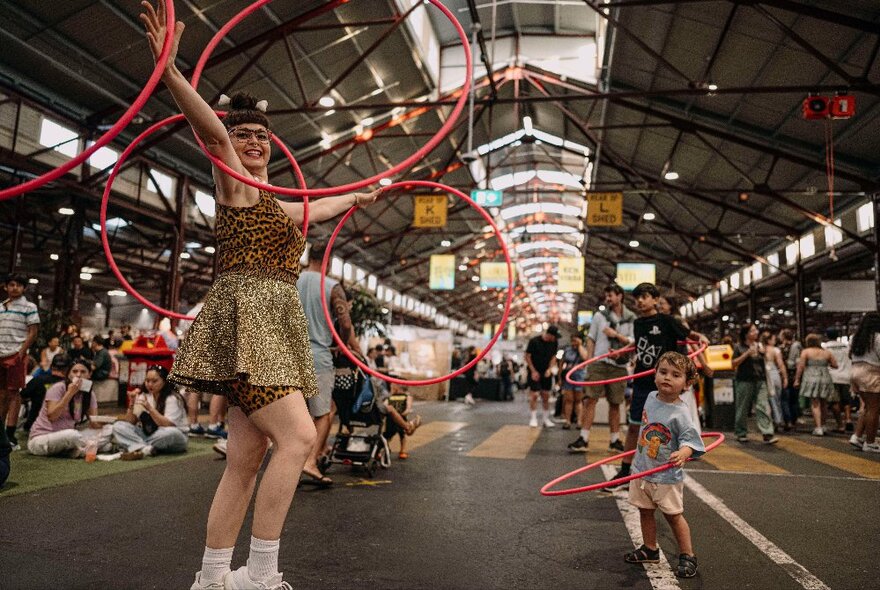 People walking around under the sheds at the Queen Victoria Summer Night market, some partaking in a hula hoop activity with an entertainer.