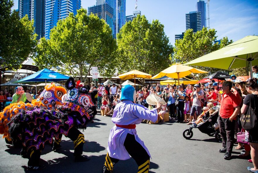 Lion dance performers dancing in front of a large crowd gathered under umbellas, with city buildings in the background.