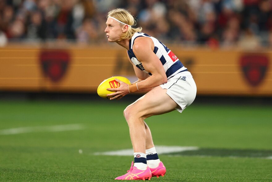 A Geelong AFL player holding a yellow football  in his hands, about to make a play, with blurred fans in the stadium in the background.