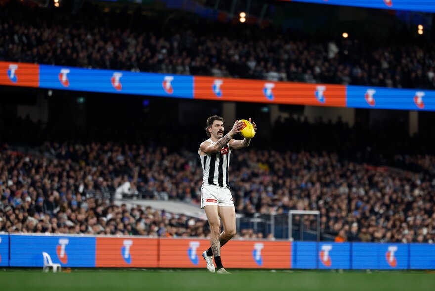 A Collingwood AFL player marking a football, with a blurred stadium crowd behind him.