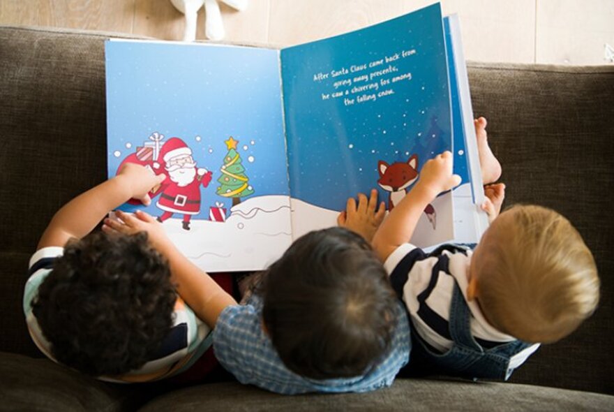 Three children reading a Santa picture book on a couch.