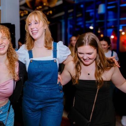 Three friends laughing and dancing with their arms around each other's shoulders, inside a restaurant.