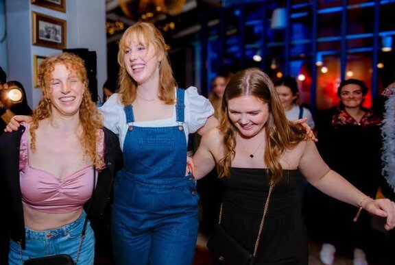 Three friends laughing and dancing with their arms around each other's shoulders, inside a restaurant.