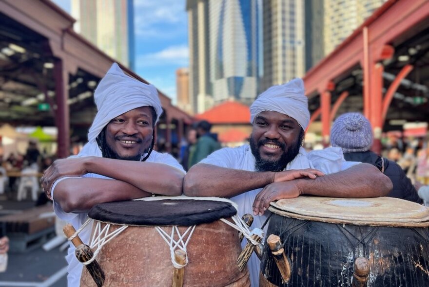 Two smiling men in white hats leaning on large African drums at Queen Victoria Market with city buildings behind them. 
