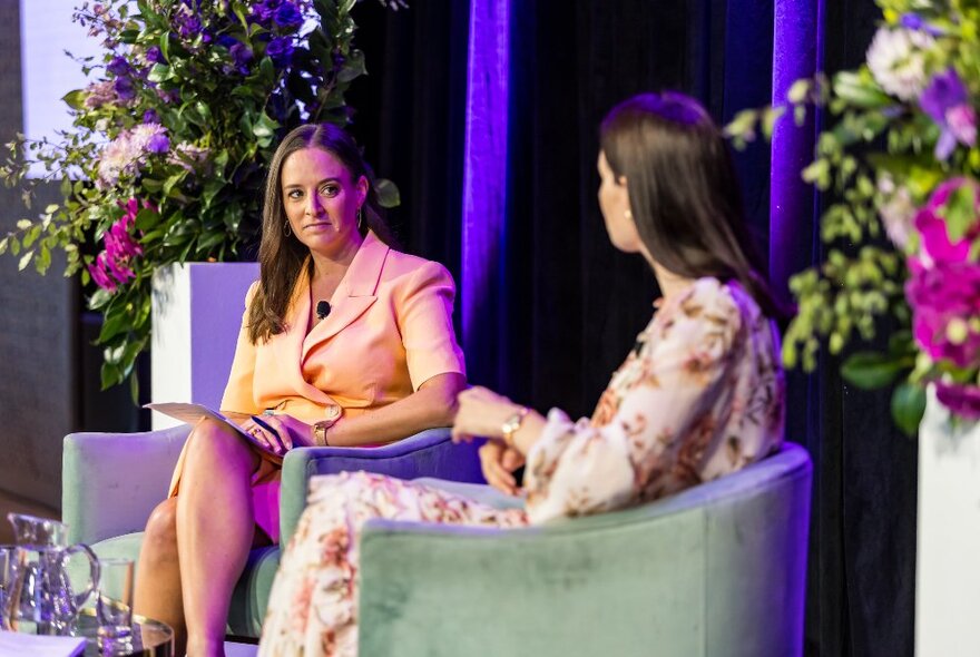 Two professional women seated in light green bucket chairs with floral arrangements behind them.