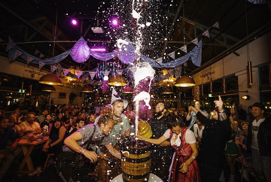 People breaking open a keg of beer, the slashes rising in the air in a room of people.