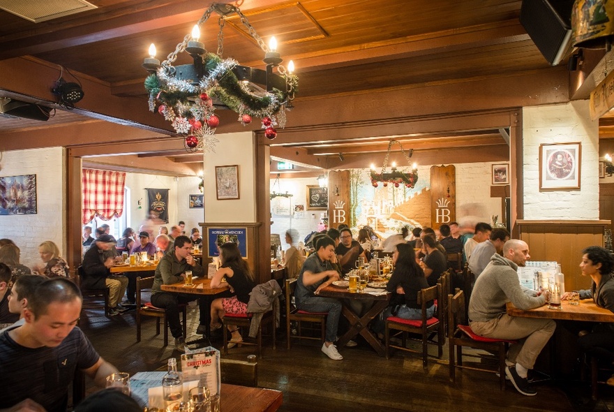 Large dining room in a restaurant with wood panelled walls and diners eating and drinking at tables, Christmas decorations wrapped around the large light hanging from the ceiling.