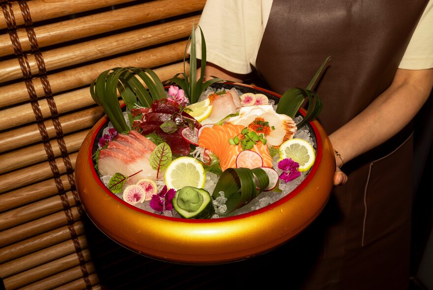 A waiter holds a gold bowl of sashimi presented on ice with decorative garnishes of flowers and leaves. 