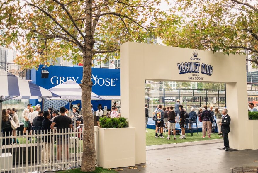 The concrete arched entrance to the Grey Goose Racquet Club, with patrons standing on synthetic turf under the shade of trees.