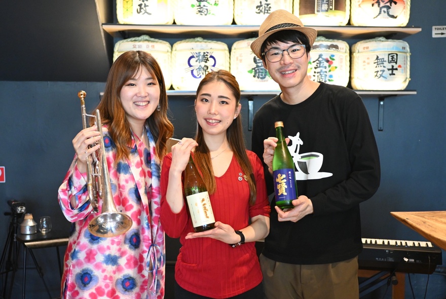 Two people holding Japanese sake bottles and a third holding a trumpet, in a Japanese bar setting in Melbourne.