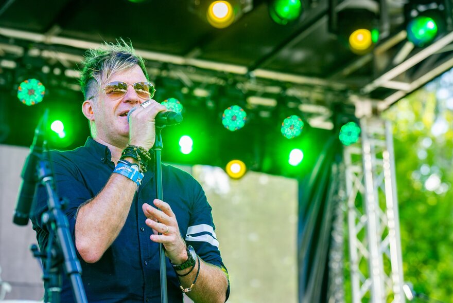 A singer wearing aviator sunglasses performing on an outdoor stage with green lights behind him.