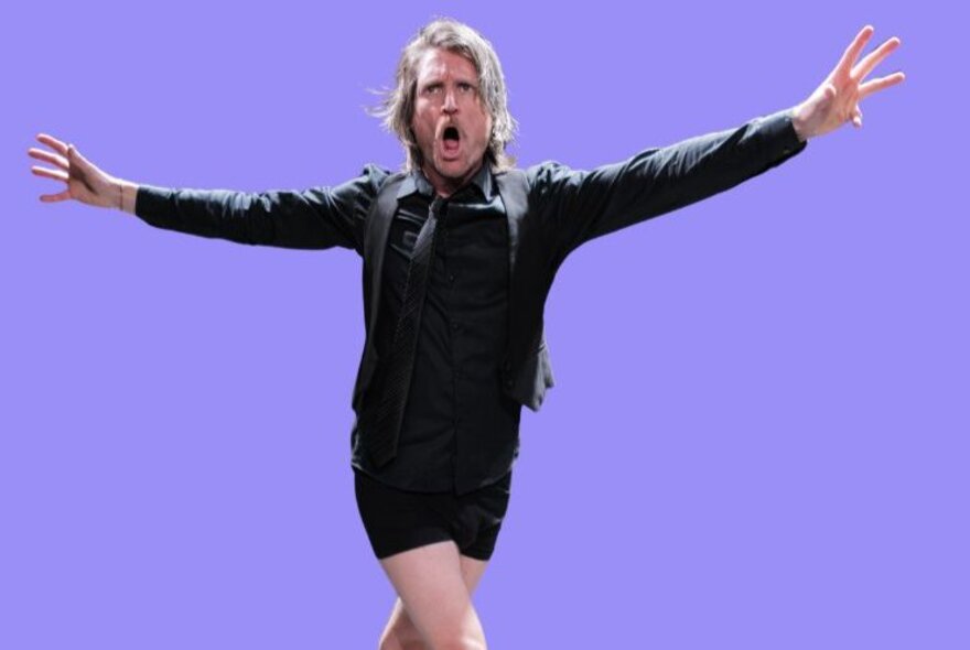 A full-shot image captures a man with longish blonde hair performing dynamically on a solid purple background.