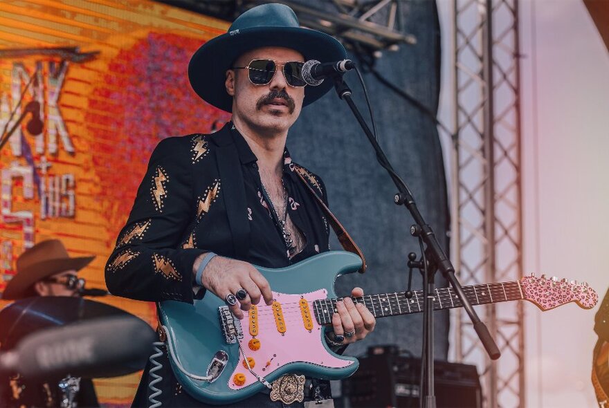 A guitarist on stage at a concert with a hat, sunglasses and lightening bolts on his shirt. 