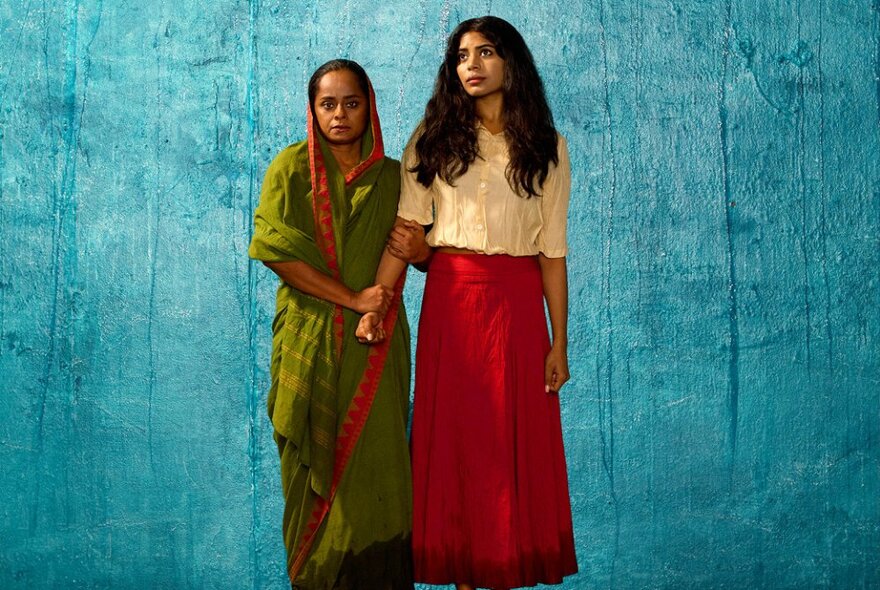 Two actors, one in Western dress, the other in traditional Indian robes, against a turquoise background.