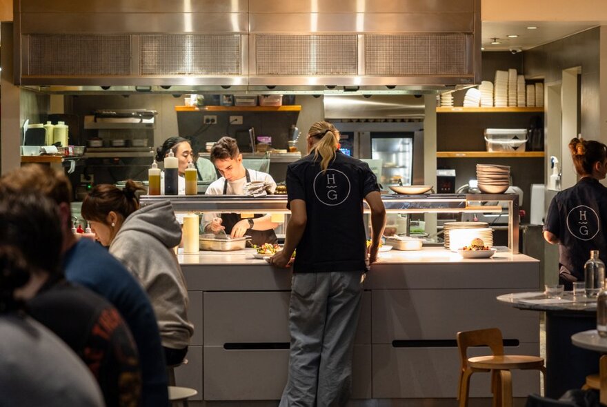 Staff serving food at a pass in an industrial-style restaurant kitchen with diners perched on stools.