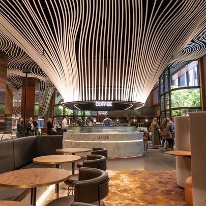 An indoor architectural space featuring a central modern coffee bar with a striking ceiling installation made of numerous curved, flowing white lines that converge above the counter, with casual seating in the foreground.