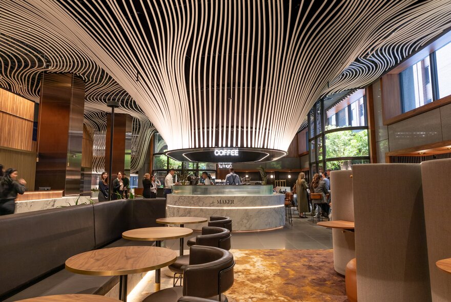 An indoor architectural space featuring a central modern coffee bar with a striking ceiling installation made of numerous curved, flowing white lines that converge above the counter, with casual seating in the foreground.