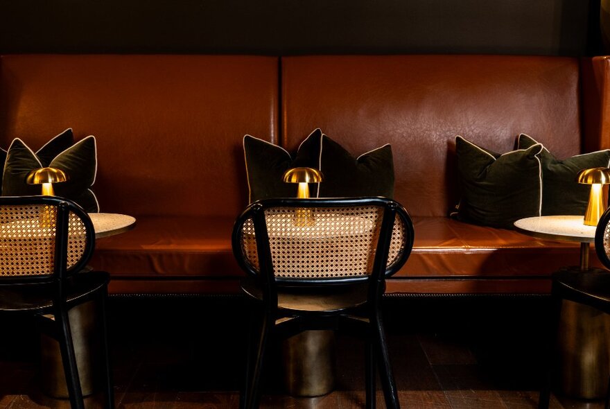 A leather bench seat with velvet cushions and small round tables with black and rattan bentwood chairs in front of it.