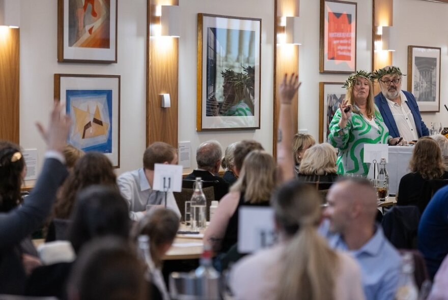 A room of people sitting at tables participating in a quiz, the trivia hosts standing behind a lectern both wearing laurel wreaths on their heads.