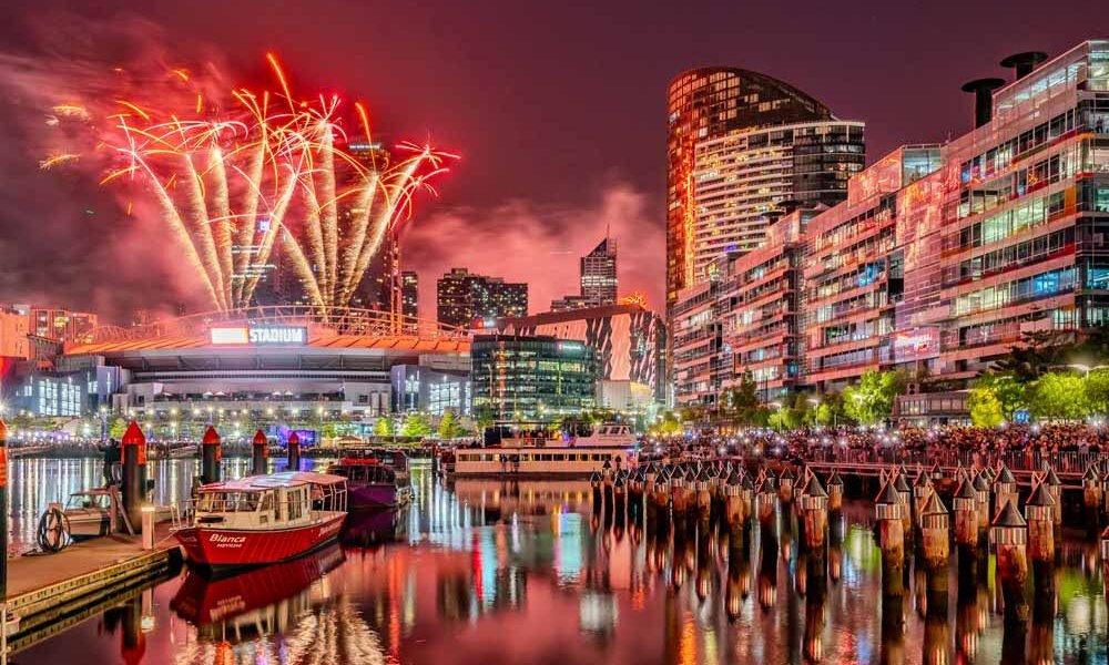 A city harbour at night, with skyscrapers and fireworks in the background. 