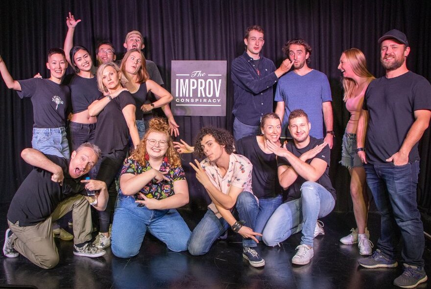 Group of people in two rows against black stage curtain, laughing and posing; Improv sign at centre.