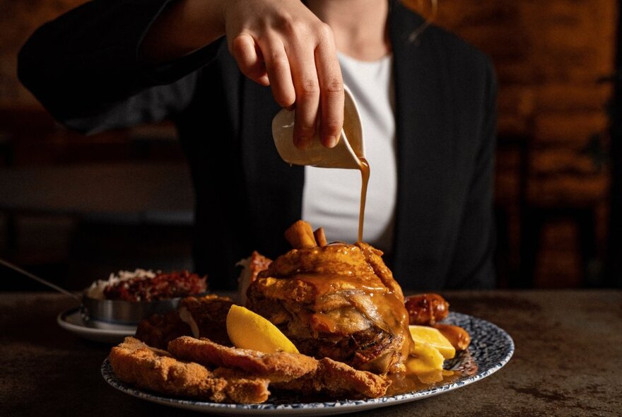 A hand pouring sauce over a plate of meat and potatoes.