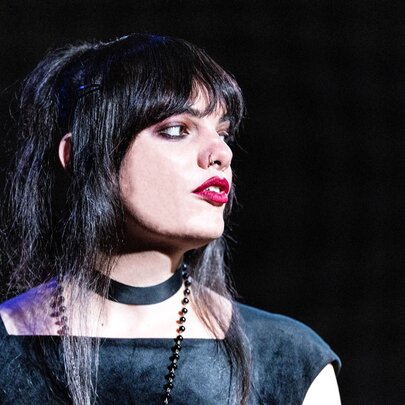 Australian comedian, writer, and performer Cassie Workman, on stage in profile, with dark hair and a fringe, wearing a choker necklace and a black top. 