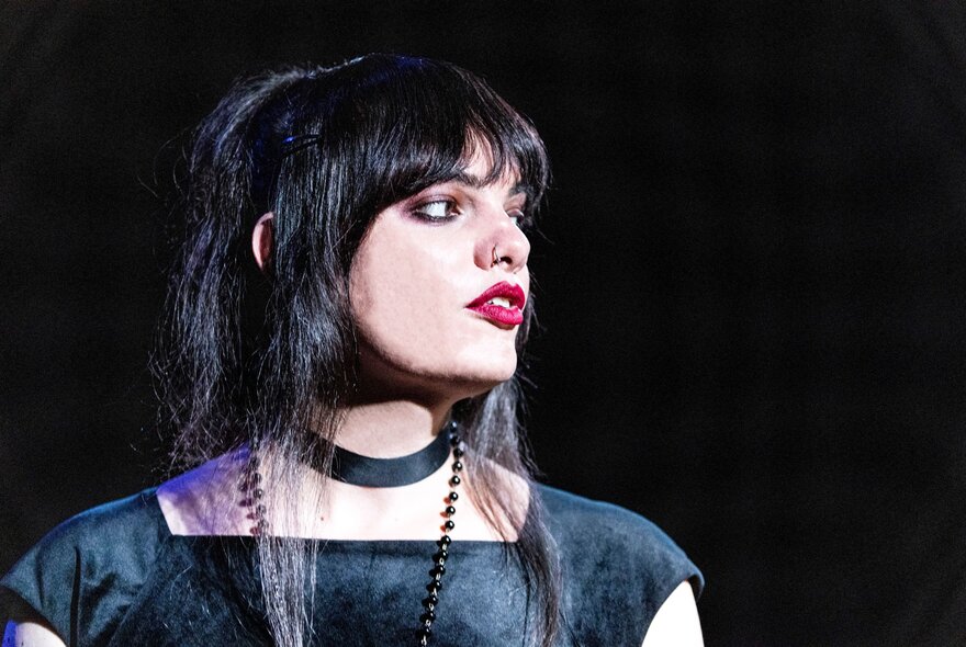 Australian comedian, writer, and performer Cassie Workman, on stage in profile, with dark hair and a fringe, wearing a choker necklace and a black top. 