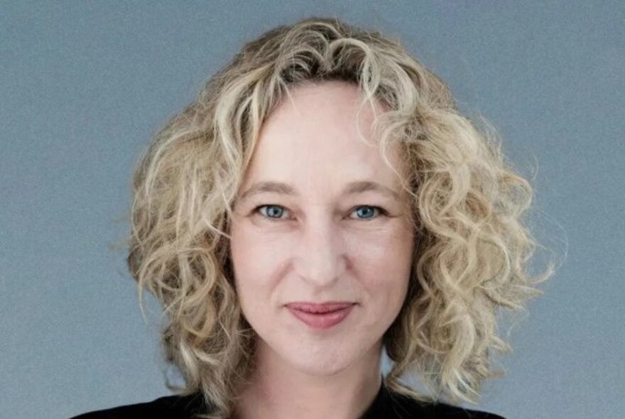 Portrait of Melbourne broadcaster, Sally Warhaft, with curly blonde hair against a blue background. 