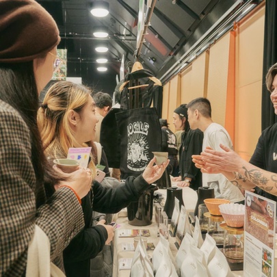 Tattooed barista handing a tasting cup to a customer next to bags of beans.
