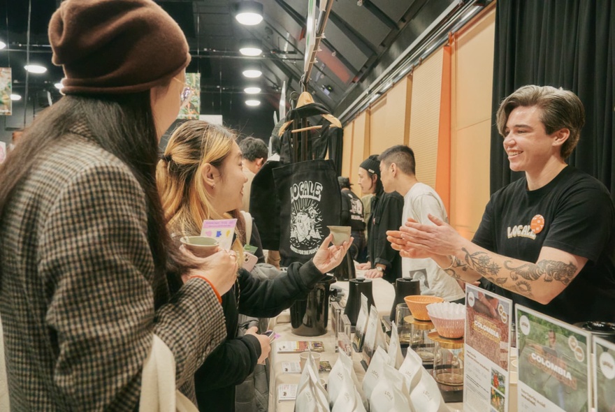 Tattooed barista handing a tasting cup to a customer next to bags of beans.