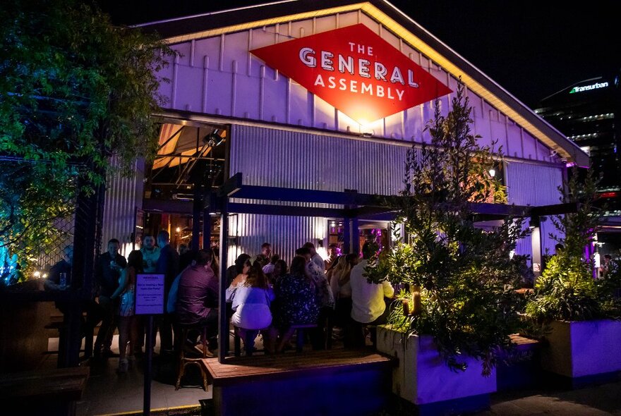 Boatshed pub exterior at night with crowd mingling around trees.