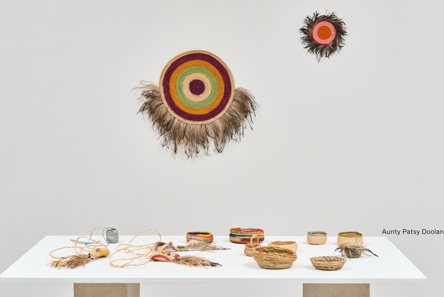 Textiles and woven objects hanging on a white wall, with woven objects on a low table display, in the white gallery space at Craft Victoria.