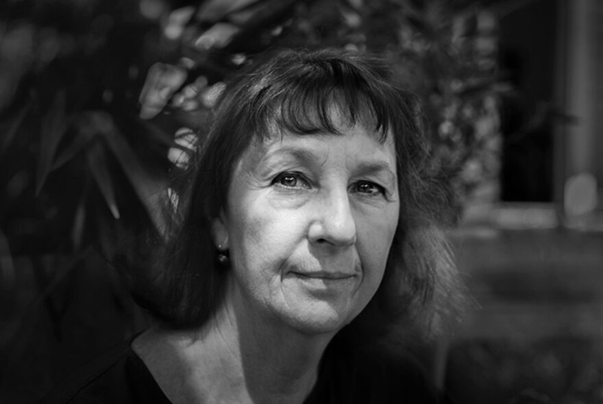 A black and white portrait of artist, Rosalie Ham, looking directly at the camera, leafy bushes behind her.
