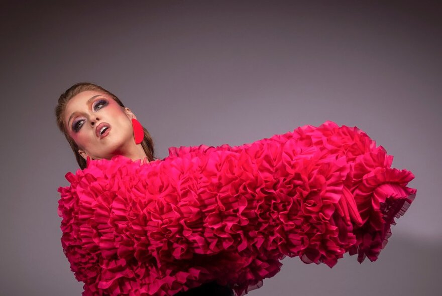 Performer wearing bright pink ruffled outfit with pink make-up and earrings, her arms raised in front of her and her head back.