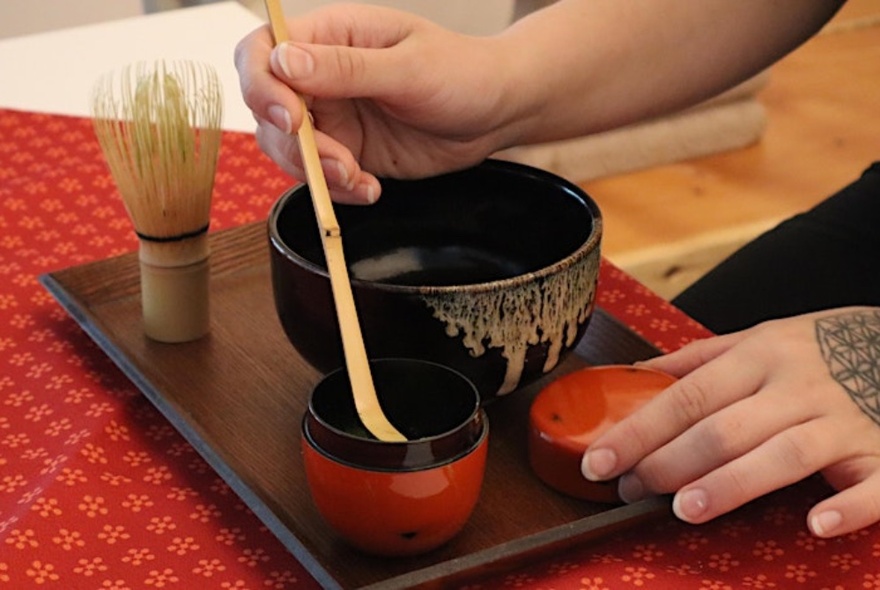 A hand whisking a Japanese tea in traditional Japanese dishes on a wooden tray, with a traditional matcha whisk.