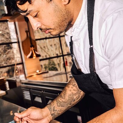 A tattooed chef plating food in a restaurant kitchen.