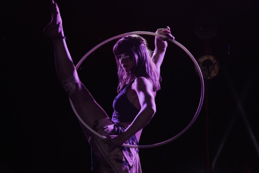 A dancer putting her leg through a hoop with a purple light cast on her. 